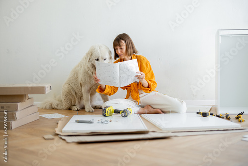 Young woman assembles furniture by herself, sitting with dog and reading manual at new apartment. DIY and house improvement concept photo