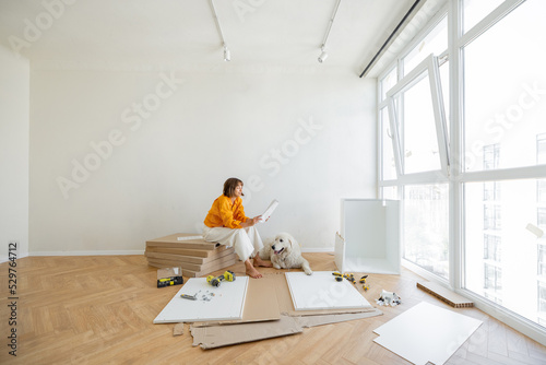 Young woman assembles furniture by herself, sitting with dog near window and reading manual at new apartment. Wide view with copy space photo