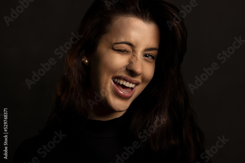 Headshot of beautiful young woman in black dress and gray background. She smile and wink on one eye