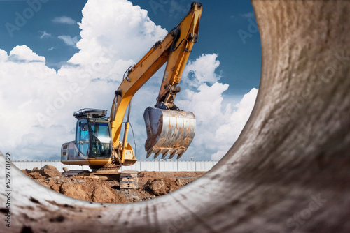 A powerful caterpillar excavator digs the ground against the blue sky. Earthworks with heavy equipment at the construction site. View from a large concrete pipe.