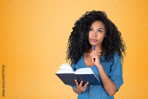 Black woman with notebook in hands, thinking portrait on empty yellow background