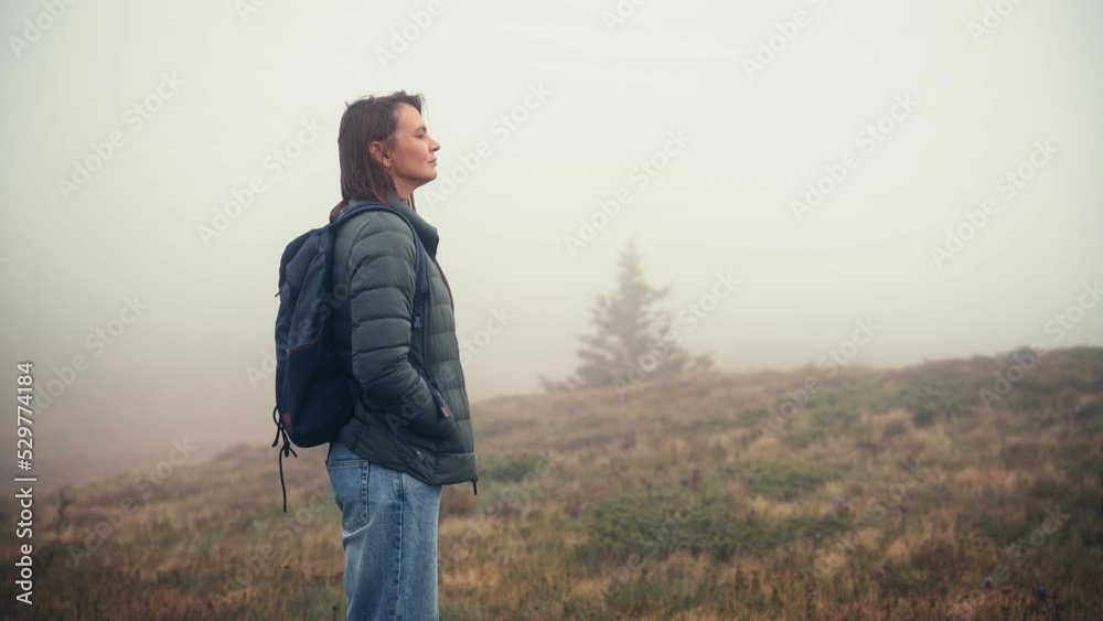 A caucasian woman with a backpack enjoying the fresh air while standing in the mountain meadow covered with thick fog. A caucasian woman with a backpack enjoying the fresh air while standing in the mountain meadow covered with thick fog.
