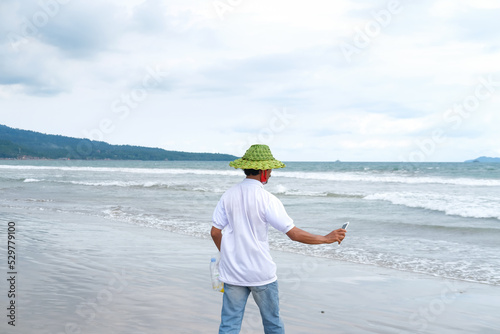 A man in a hat made of woven young coconut leaves walks on the beach holding a smartphone