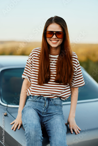 A fashionable woman smiles sweetly in stylish sunglasses, a striped t-shirt and jeans, sits on the trunk of a car and looks at the beautiful nature of autumn. Travel lifestyle © SHOTPRIME STUDIO