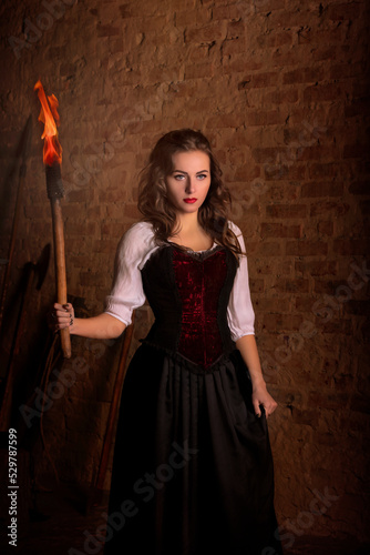 Fantastic medieval woman with a torch in her hands. Royal velvet vintage red dress with fur and gold, princess, queen, brunette with long hair. Castle, dusk, night, luxury