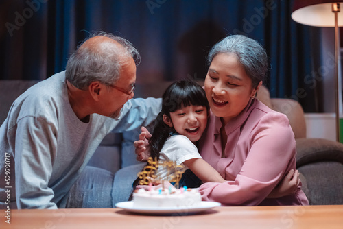 Asian happy family of little girl hugging lover with grandparents after blowing out candles on cake. Celebrate birthday anniversary party on table at night in living room  having happiness lifestyle.