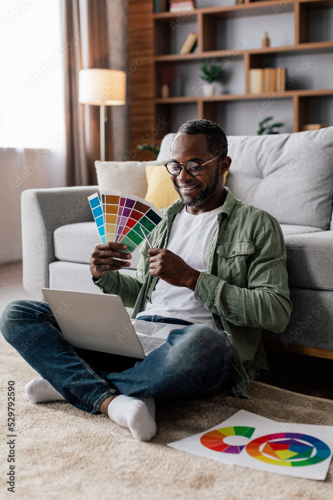 Fototapeta premium Cheerful adult black guy designer in glasses and casual show color palettes in laptop on floor in living room