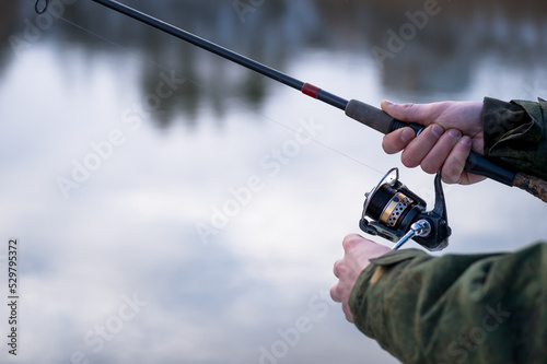 The frozen hands of a male angler close-up to hold the spinning in a blurry motion wind the fishing line on a reel against the background of a winter river. Selective focus on the hand. Background