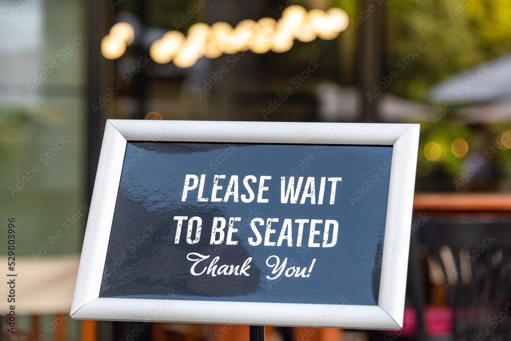 Please wait to be seated sign standing at the front of a restaurant. Sidewalk cafe hostess stand with message signboard for clients.