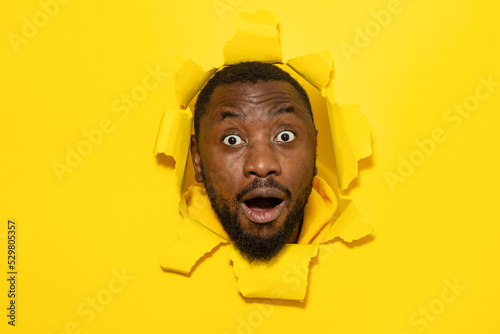 Shocked african american man reacting with surprisement, looking at camera, posing in torn yellow paper hole