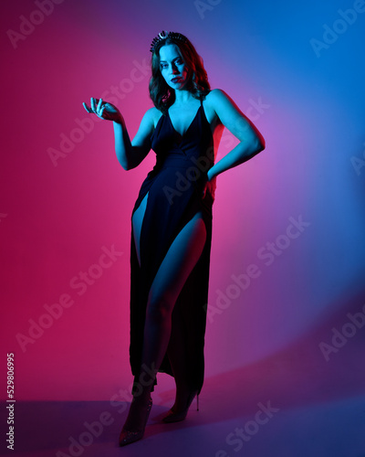 Close up portrait of beautiful woman model wearing elegant black dress and crown, posing against a studio background with fantasy inspired arm gestures, multi coloured creative lighting.