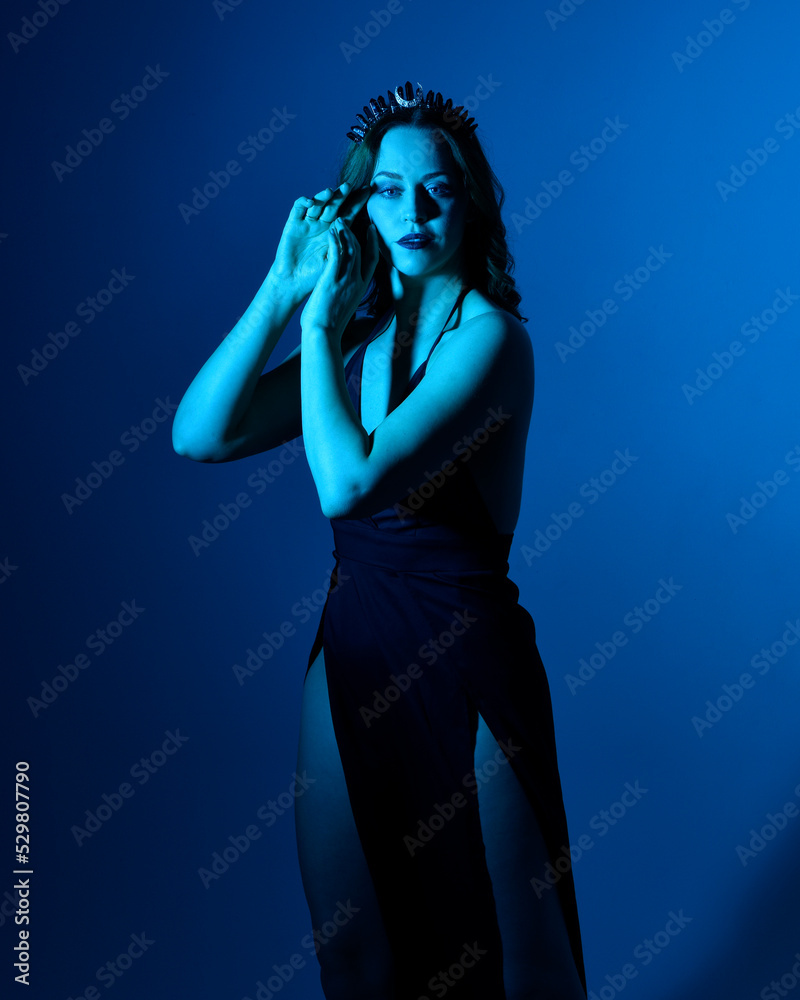 Close up portrait of beautiful woman model wearing elegant black dress and crown, posing against a studio background with fantasy inspired arm gestures, multi coloured creative lighting. Close up portrait of beautiful woman model wearing elegant black dress and crown, posing against a studio background with fantasy inspired arm gestures, multi coloured creative lighting.