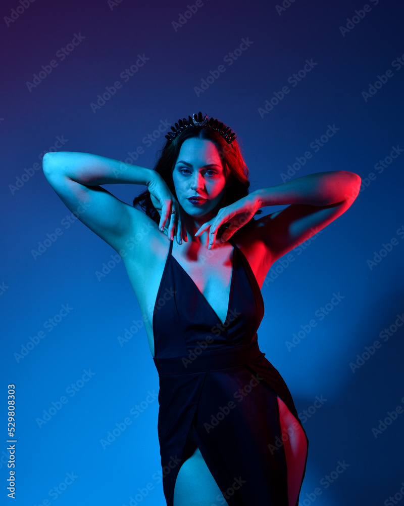 Close up portrait of beautiful woman model wearing elegant black dress and crown, posing against a studio background with fantasy inspired arm gestures, multi coloured creative lighting.
