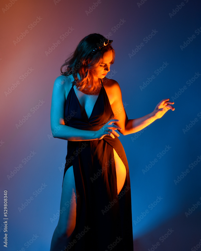 Close up portrait of beautiful woman model wearing elegant black dress and crown, posing against a studio background with fantasy inspired arm gestures, multi coloured creative lighting.
