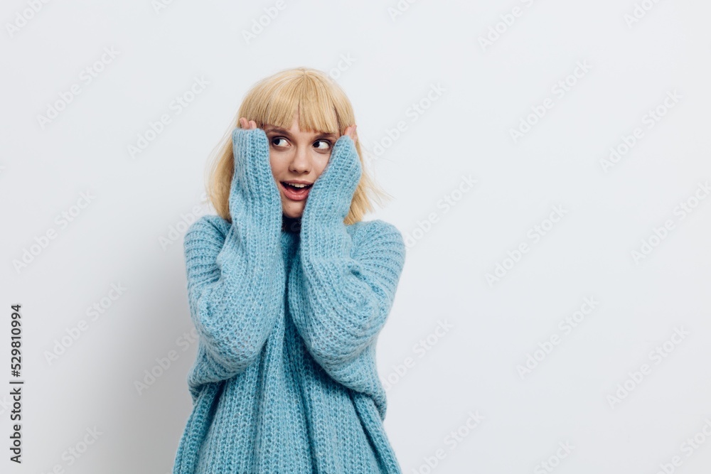 a happy, joyful woman stands on a light background in a blue sweater and looks up in surprise with her hands on her face a happy, joyful woman stands on a light background in a blue sweater and looks up in surprise with her hands on her face