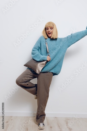 a full-length photo of a beautiful, happy, carefree stylish woman in a blue sweater and with a large bag, vigorously posing with her hand to the side