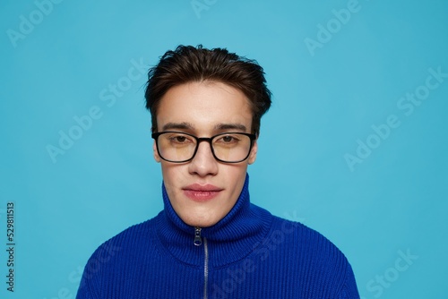 horizontal portrait of a handsome man in a blue zip-up sweater and black eyeglasses, standing on a light blue background, looking pleasantly into the camera