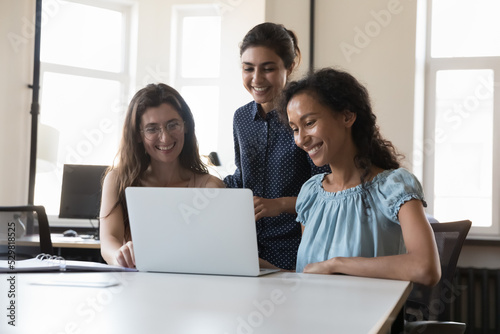 Happy successful diverse team of three employees women using laptop together at shared workplace, looking at monitor, laughing, talking on video conference call, enjoying online communication © fizkes