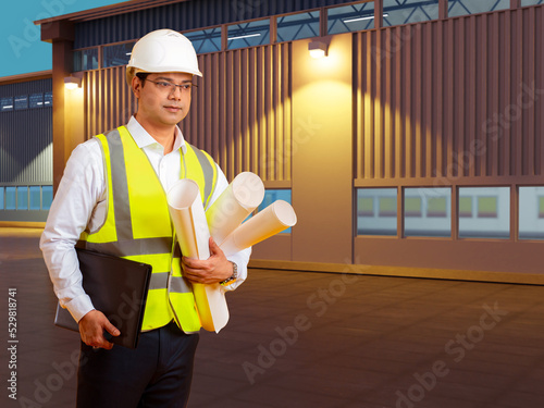 Man engineer. Builder next to new factory building. Guy with blueprints in evening industrial zone. Concept construction of plants and factories. Employee engineering firm. Factory engineer career photo