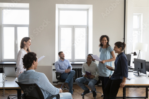 Happy office friends chatting in office, discussing success, sharing ideas, brainstorming on project, laughing, smiling. Diverse team meeting at workplaces, sitting, standing in circle