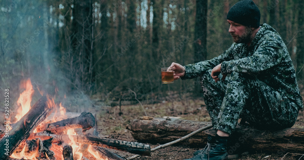Man with cup of tea the fire in the forest. Human looks at the bonfire and drinks tea on the nature in the summer. Alone traveler sits and rests by the fire with a cup of coffee at dusk. Man with cup of tea the fire in the forest. Human looks at the bonfire and drinks tea on the nature in the summer. Alone traveler sits and rests by the fire with a cup of coffee at dusk.