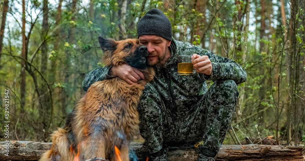 Adult Man is relaxing at the fire with his dog in the nature. Alone traveler is resting with his German shepherd by the forest fire and drinking tea. Man strokes a dog at the fire in the forest.