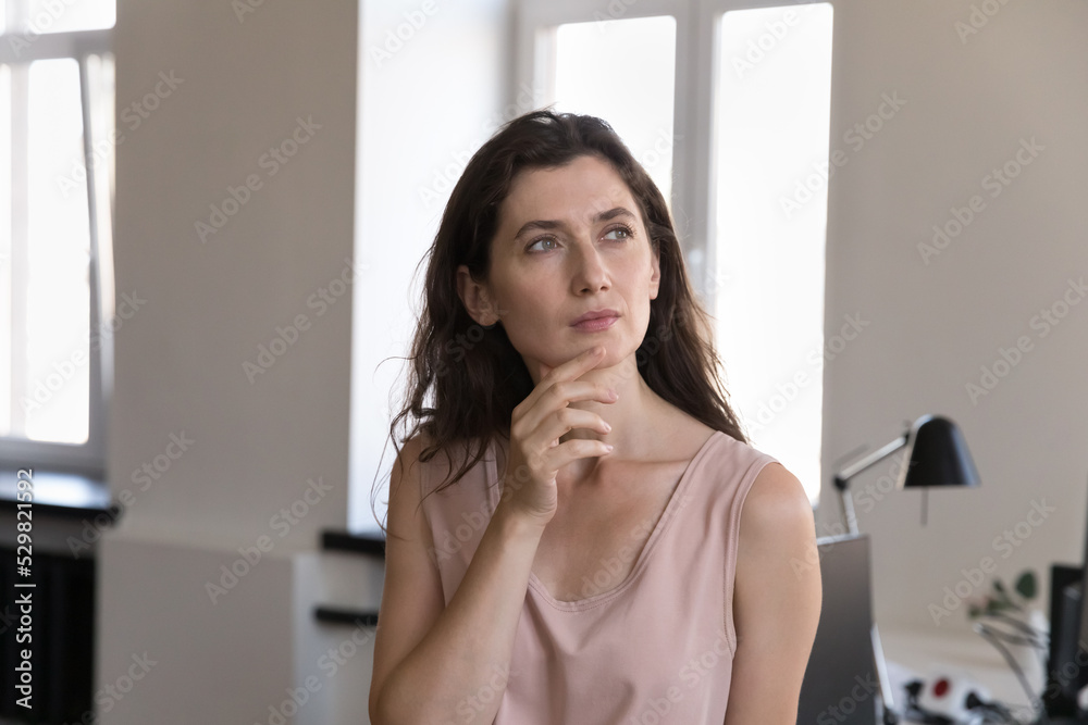 Serious young Caucasian business woman in casual standing, posing at office workplace, touching chin, looking away with thoughtful face, dreaming, thinking over project tasks, problem solving Serious young Caucasian business woman in casual standing, posing at office workplace, touching chin, looking away with thoughtful face, dreaming, thinking over project tasks, problem solving