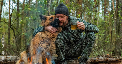 Adult Man is relaxing at the fire with his dog in the nature. Alone traveler is resting with his German shepherd by the forest fire and drinking tea. Man strokes a dog at the fire in the forest.