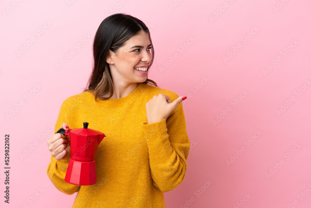 Young Italian woman holding a coffee maker isolated on pink background pointing to the side to present a product Young Italian woman holding a coffee maker isolated on pink background pointing to the side to present a product