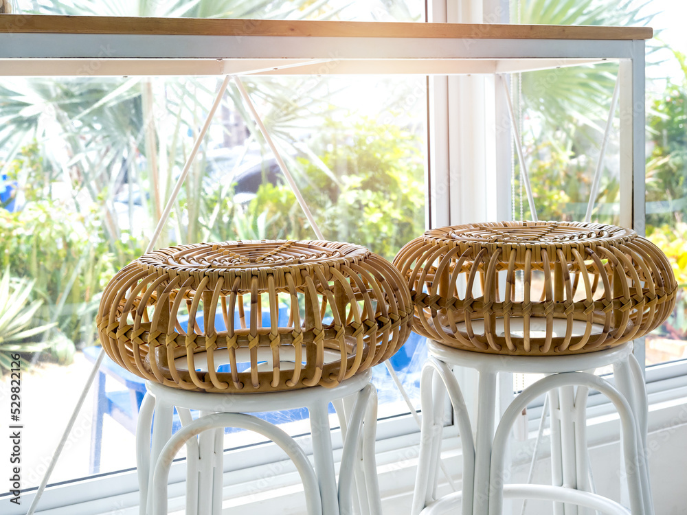 Two empty round rattan bar stool chairs near the table and glass window decoration in cafe with green tropical palm tree view outside.