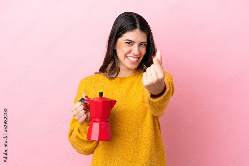 Young Italian woman holding a coffee maker isolated on pink background making money gesture Young Italian woman holding a coffee maker isolated on pink background making money gesture
