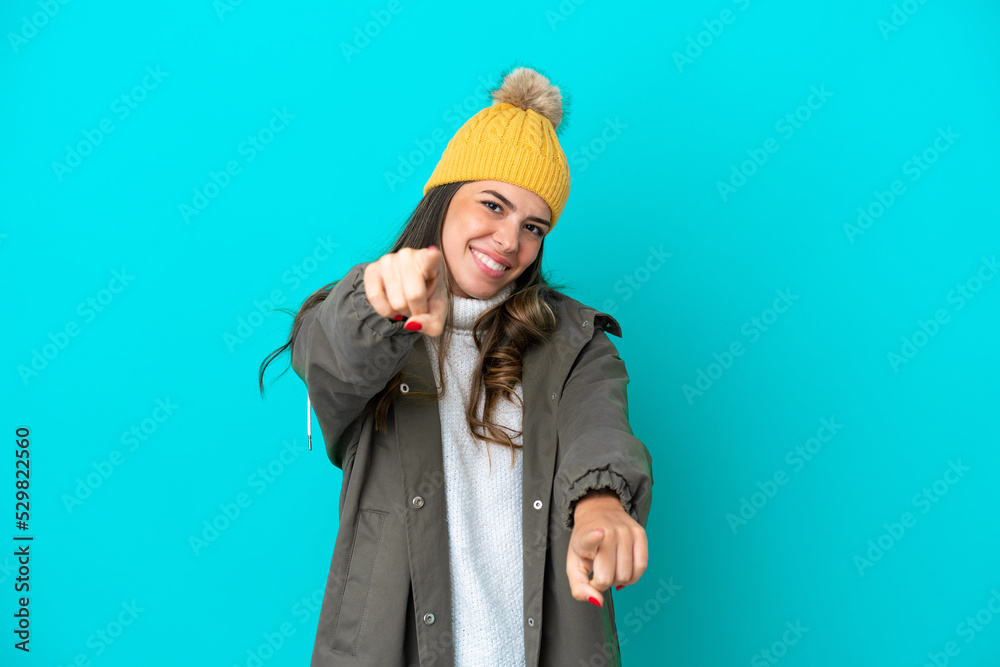 Young Italian woman wearing winter jacket and hat isolated on blue background points finger at you while smiling Young Italian woman wearing winter jacket and hat isolated on blue background points finger at you while smiling
