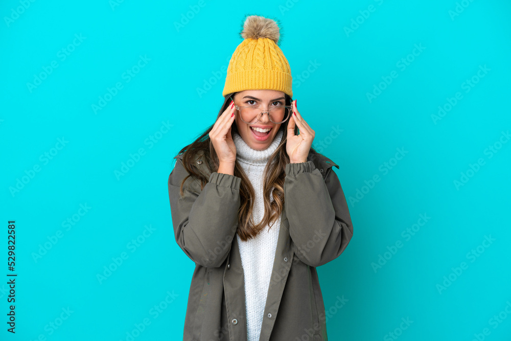 Young Italian woman wearing winter jacket and hat isolated on blue background with glasses and surprised Young Italian woman wearing winter jacket and hat isolated on blue background with glasses and surprised