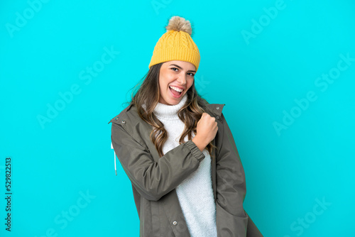 Young Italian woman wearing winter jacket and hat isolated on blue background celebrating a victory