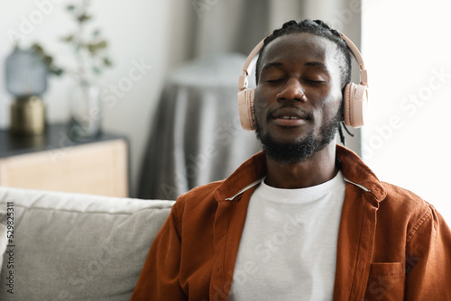 Calm african american man in wireless headphones sitting on sofa with closed eyes and listening to music, free space
