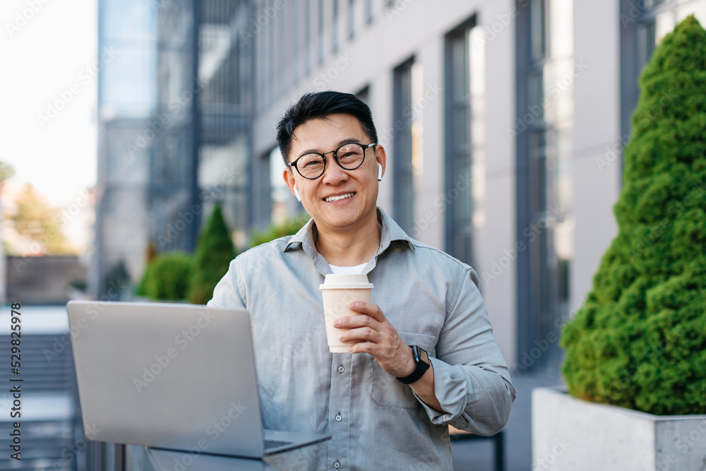 Happy asian middle aged businessman working on laptop and drinking coffee outdoors near office building Happy asian middle aged businessman working on laptop and drinking coffee outdoors near office building