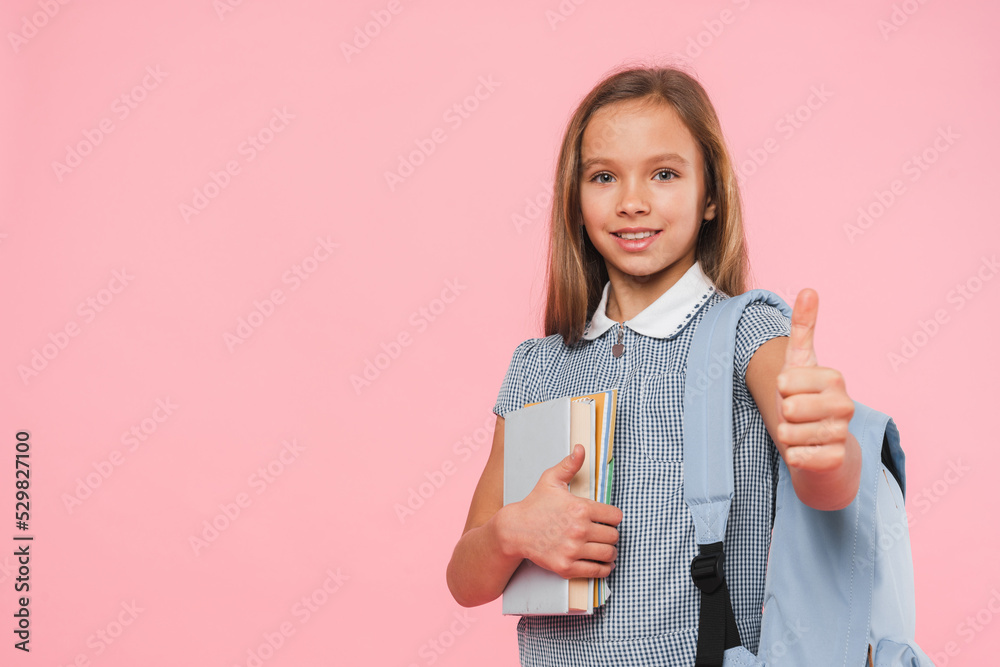 Closeup photo of young caucasian preteen schoolgirl teenager in uniform with bag and textbooks looking at camera showing thumb up isolated in pink Closeup photo of young caucasian preteen schoolgirl teenager in uniform with bag and textbooks looking at camera showing thumb up isolated in pink