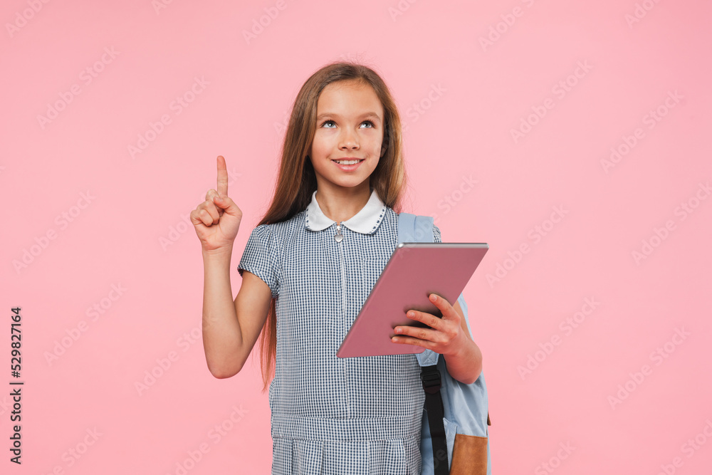 Smart teenage preteen schoolgirl in uniform holding digital tablet for e-learning remote education online lessons having idea isolated in pink background