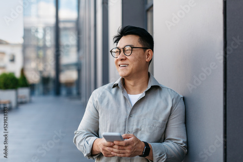 Confident korean middle aged businessman holding smartphone and looking aside at free space, standing outdoors photo