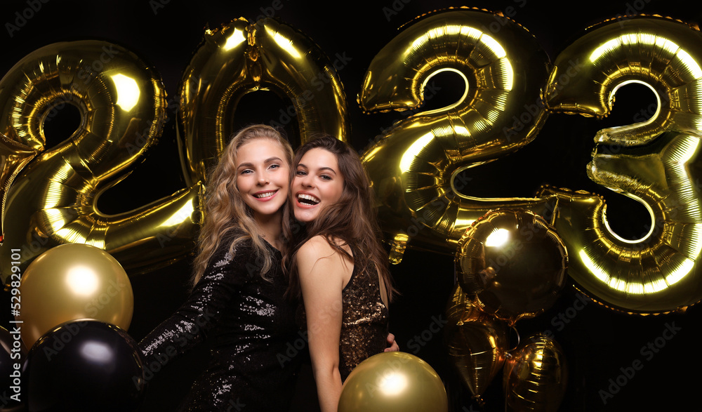 Happy gorgeous girls in stylish sexy party dresses holding gold 2023 balloons, having fun at New Year's Eve Party. - obrazy, fototapety, plakaty 