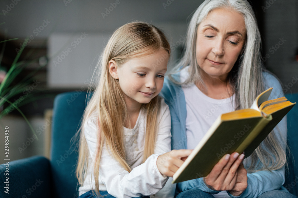 Serious caucasian small granddaughter and mature grandmother are reading book on sofa in room interior Serious caucasian small granddaughter and mature grandmother are reading book on sofa in room interior