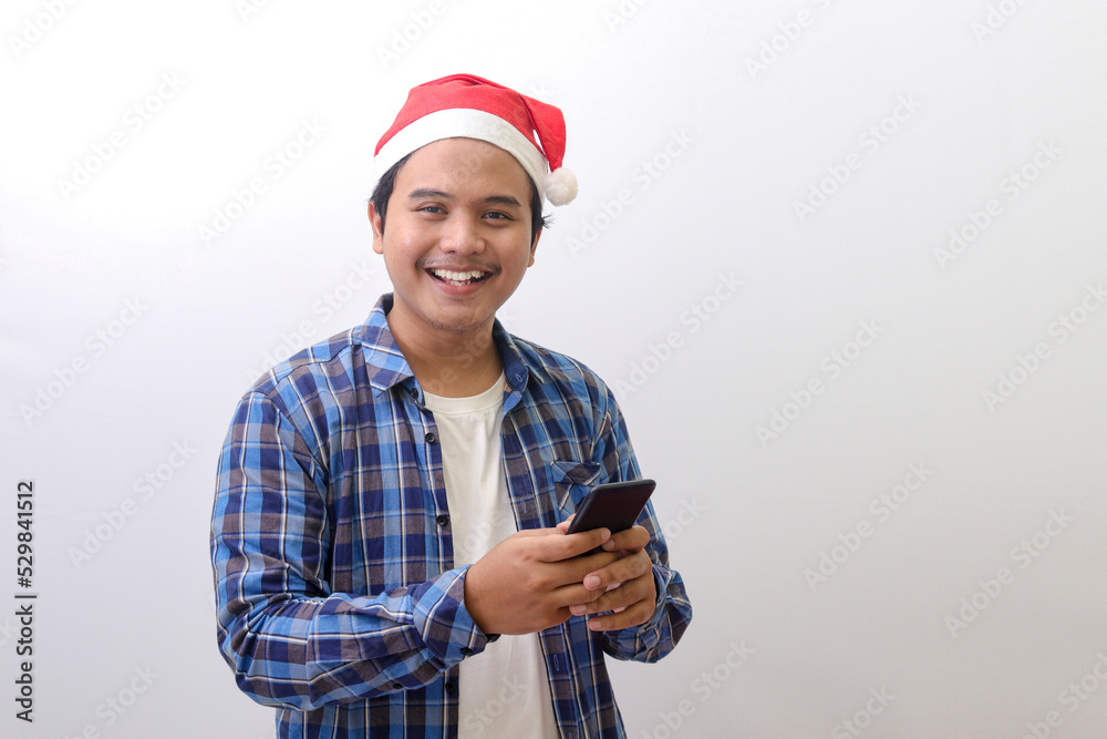 Portrait of excited Asian man in blue plaid shirt wearing santa hat looking at camera and holding a mobile phone. Advertising concept. Isolated image on gray background Portrait of excited Asian man in blue plaid shirt wearing santa hat looking at camera and holding a mobile phone. Advertising concept. Isolated image on gray background
