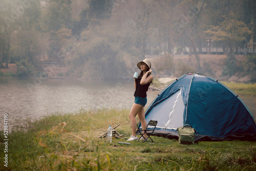 Happy woman drinking coffee in front of the camping tent at meadow near lake. Recreation and journey outdoor activity lifestyle. Travel and adventure theme.