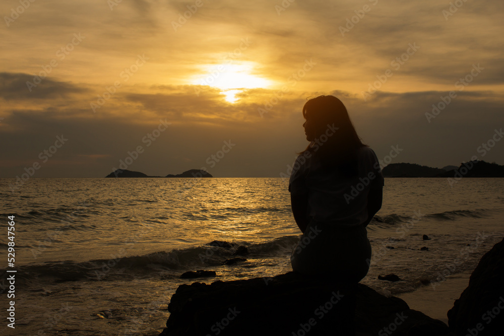 Silhouette of long hair woman sit on the stone beside the beach and wave at sunset sunrise