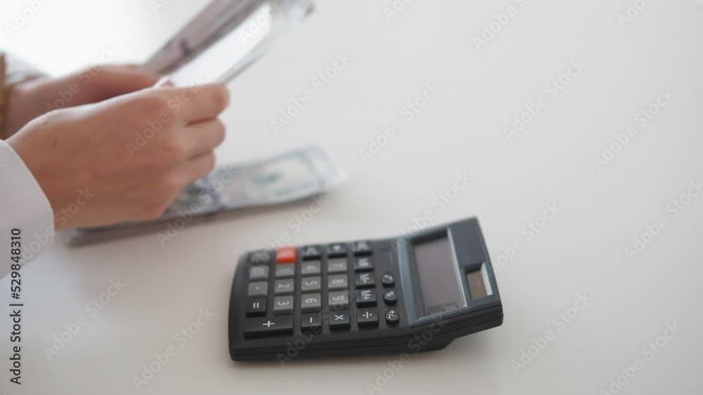 Budget spending. Busy woman. Money counting. Unrecognizable lady using calculator sitting desk with cash dollars in light room interior. Budget spending. Busy woman. Money counting. Unrecognizable lady using calculator sitting desk with cash dollars in light room interior.