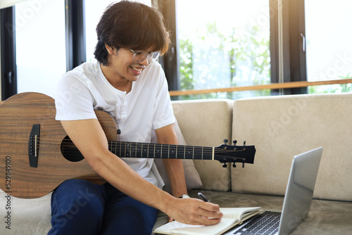 Fotografie An Asian man in a white shirt writes down ideas from his acoustic guitar in a notebook as a weekend songwriting staple in his own home