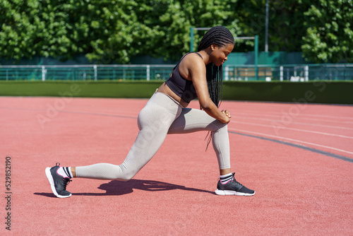 Sportive young woman with long braids takes care of body on summer day. African American lady in sportswear enjoys effective exercise for gluteal muscles on city stadium near park with green trees