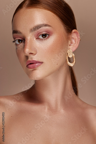 Female model with red hair, brown eyes, freckled skin, poses for a beauty shoot. She wears gold earrings and has her hand to her face.  photo