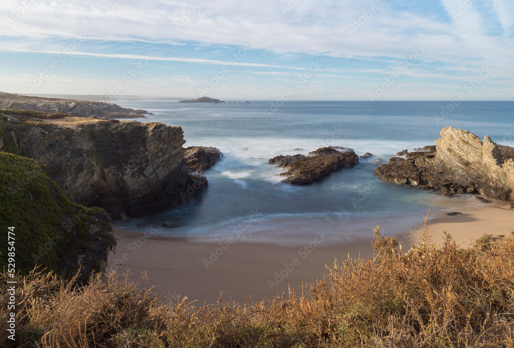 View of empty Praia dos Buizinhos sand beach with long exposure blurred ocean waves and sharp rock and cllifs at wild Vicentina coast in Porto Covo, Portugal. Golden hour.