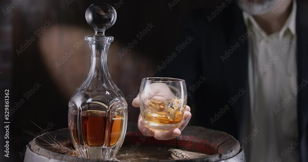 Man in a suit and beard (sommelier) takes a glass of whiskey on ice from a vintage whiskey barrel and shakes the glass to spread the smell of whiskey. Slow motion 4K